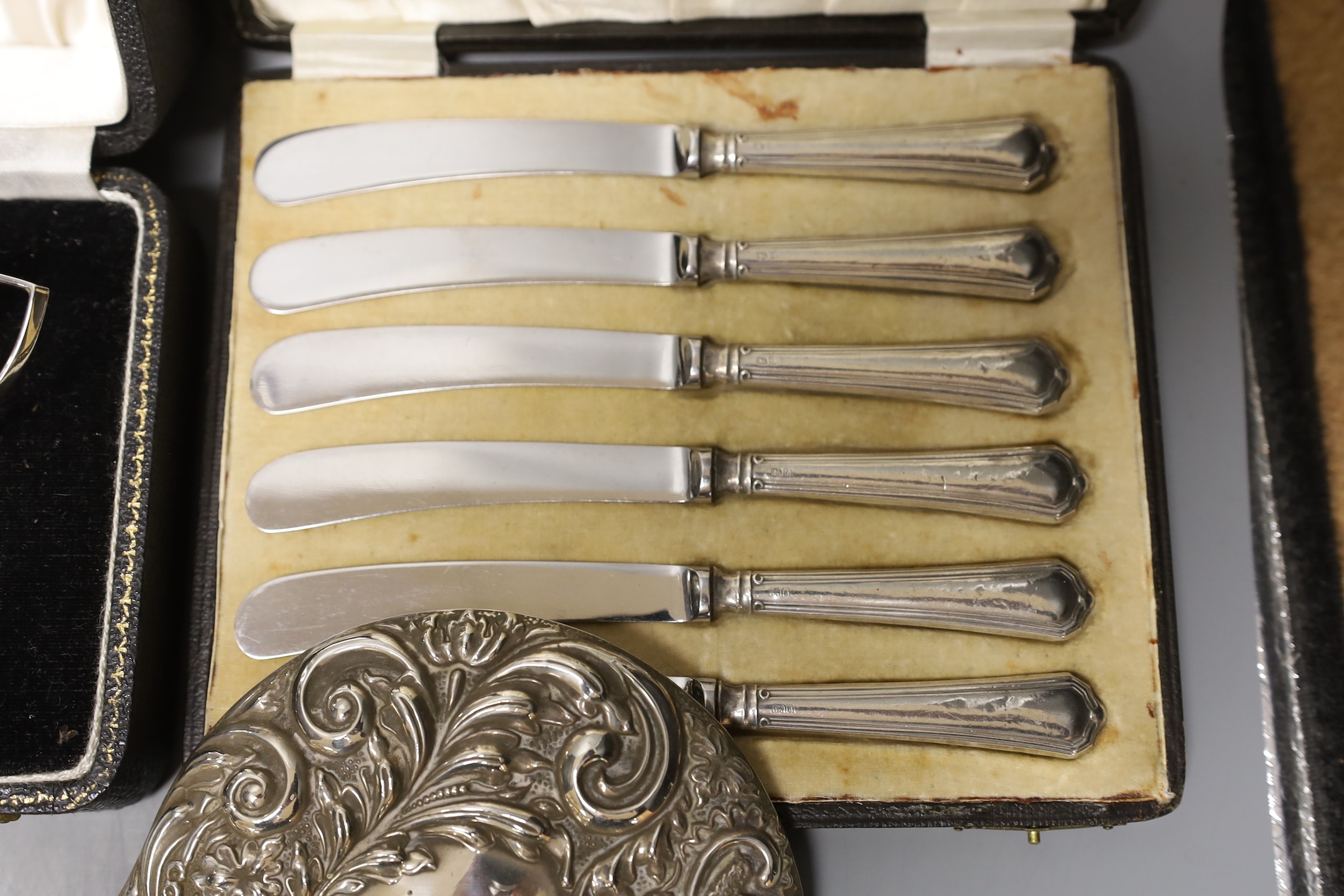 A cased 1950's silver three piece condiment set, two cased sets of six silver handled tea knives, a silver mounted glass salts bottle, silver mounted hand mirror and two brushes.